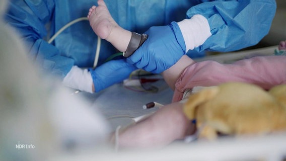 Eine Krankenpflegerin oder ein Krankenpfleger versorgt ein Kleinkind, von dem man nur den liegenden Unterkörper und einen Fuß sieht. © Screenshot 