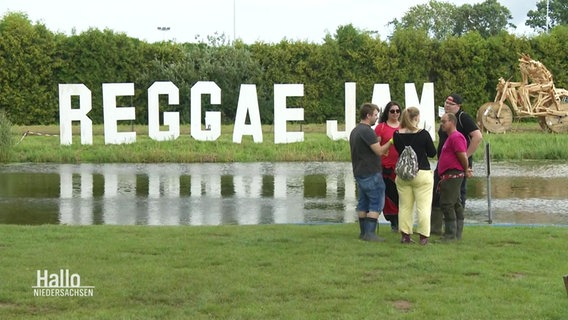 Große weiße Buchstaben im Hintergrund bilden den Schriftzug "REGGAE JAM", im VOrdergrund eine große Pütze auf einer Wiese und Menschen in Regenkleidung. © Screenshot 