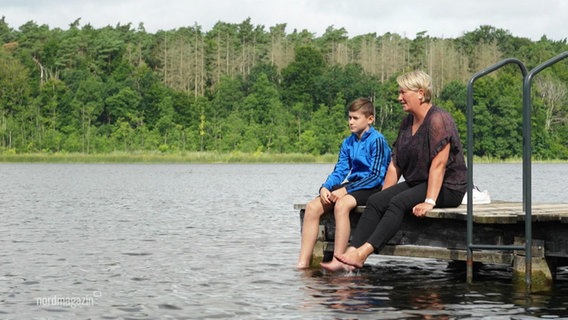 Ein Junge und eine Frau sitzen auf einem Steg und lassen ihre Füße ins Wasser hängen. © Screenshot 