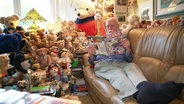 Hans-Dieter Gau sitzt mit ausgestreckten Beinen auf seiner Couch und liest eine Zeitschrift. Um ihn herum sitzen und liegen zahlreiche Teddy-Bären. © Screenshot 