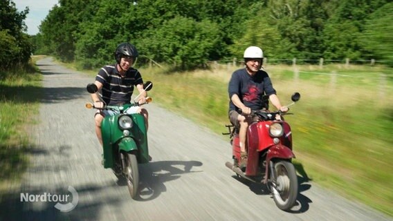 Zwei Männer mit Helmen fahren jeweils auf einer roten und einer grünen Vespa auf einer Straße nebeneinander her. Idyllische Landschaft. © Screenshot 