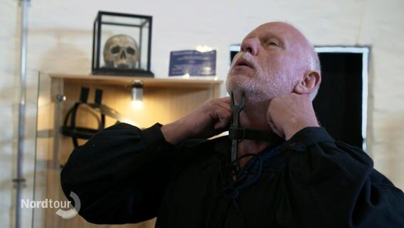 Ein Mann legt sich ein mittelalterliches Folterwerkzeug um den Hals, das ihn zwingt, den Kopf in den Nacken zu legen. © Screenshot 