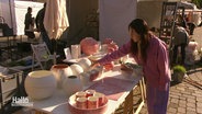 Eine Frau stellt Keramikvasen und weitere keramische Erzeugnisse auf einem Stand auf. © Screenshot 