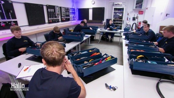 Junge Auszubildende sitzen an Tischen, vor jedem steht eine blaue Werkzeugkiste auf dem Tisch. © Screenshot 