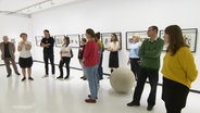 Menschen betrachten Bilder in einer Ausstellung. © Screenshot 