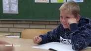 Ein Junge sitzt an einem Tisch in einem Klassenraum und stützt den Kopf mit der linken Hand, er lächelt und sieht fröhlich aus. © Screenshot 