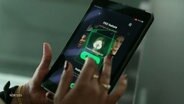Hände mit dunkelrotem Nagellack auf den Fingern tippen auf einem Smartphone, auf dessen Bildschrim Gesichter sowie ein grünes Logo zu sehen sind. © Screenshot 