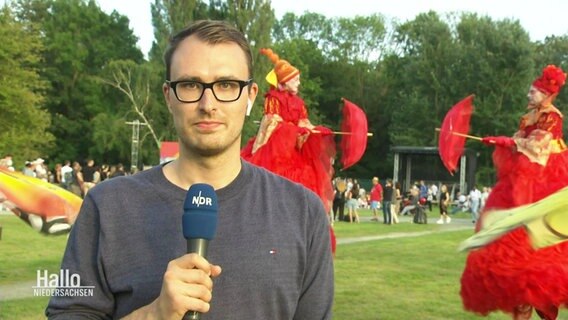 Ein NDR Moderator steht auf einer Wiese, hinter ihm sind Walking-Acts zu sehen. © Screenshot 