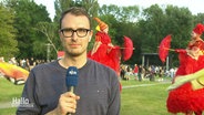 Ein NDR Moderator steht auf einer Wiese, hinter ihm sind Walking-Acts zu sehen. © Screenshot 