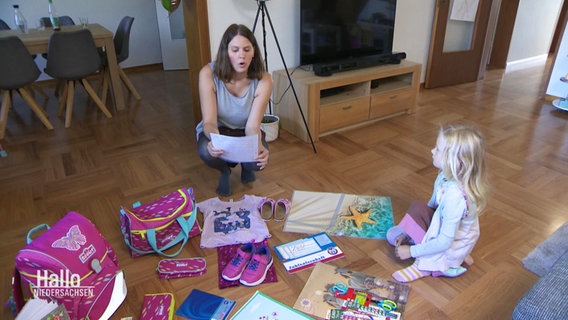 Eine Mutter sitzt mit ihrer Tochter auf dem Boden, vor ihnen eine Sammlung von Schul-Utensilien. © Screenshot 
