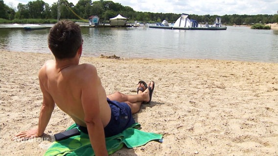 Ein Mann sitzt mit Badehose im Sand am blauen See. © Screenshot 