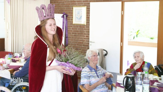 Die Heidekönigin steht in ihrem Gewand in einem Altersheim und hält einen Blumenstrauß in der Hand. © Screenshot 