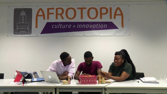 Drei junge Unternehmer:innen unterhalten sich. Hinter ihnen hängt ein Banner des Kulturzentrum "Afrotopia". © Screenshot 