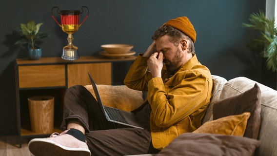 Ein Mann auf dem Sofa mit seinem Laptop auf dem Schoß schaut zerknirscht. (extra 3 vom 17.08.2023 im Ersten) © NDR 