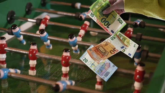 Geldscheine fliegen auf einen Tischkicker. (extra 3 vom 17.08.2023 im Ersten) © NDR 