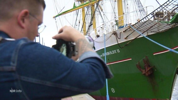 Eine Person mit Kamera vor einem Segelschiff. © Screenshot 