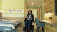 Eine Person in einem Rollstuhl fährt in ein Zimmer. © Screenshot 
