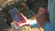 Eine Person scannt einen QR-Code auf einem Friedhof mit ihrem Smartphone. © Screenshot 