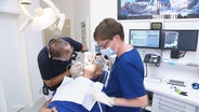 Blick in eine Zahnarztpraxis: einem Patienten wird von einem Arzt in den Mund geschaut, eine Zahnarzthelferin hält einen Schlauch. © Screenshot 