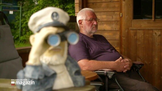 Ein Mann sitzt in einem Gartenstuhl, daneben schaut eine Figur durch ein Fernglas. © Screenshot 