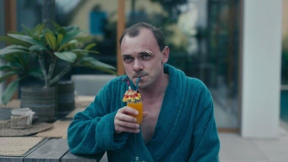 Ein Mann in blaugrünem Bademantel und geklebter Wunde auf einer Augenbraue trinkt mit einem Strohhalm einen Cocktail mit Schirmchen. © Screenshot 