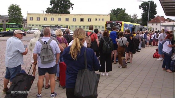 Eine lange Schlange von Menschen, die Gepäck bei sich haben, wartet vor einem Bus des Schienenersatzverkehrs. © Screenshot 