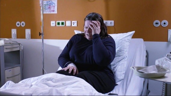 Eine Frau in einem Krankenbett fasst sich an den Kopf. © Screenshot 