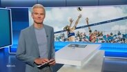 Nachrichtensprecher Thorsten Schröder, im Hintergrund ein Bild von einem Beachvolleyballspiel. © Screenshot 