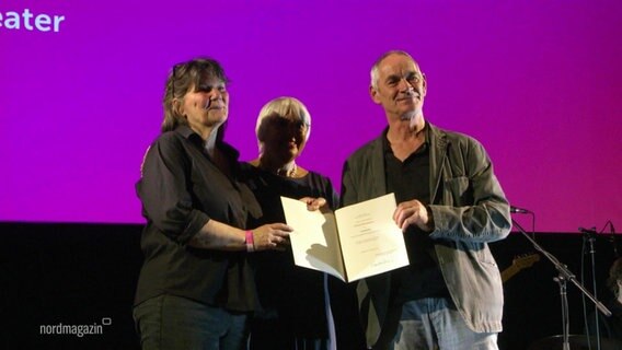 Drei Personen stehen auf einer Bühne und halten einen Preis in den Händen. © Screenshot 