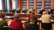 Blick in einen Gerichtssaal, über die Schulter von Menschen im Zuschauerraum, die auf die Richterbank im Vordergrund schauen. © Screenshot 