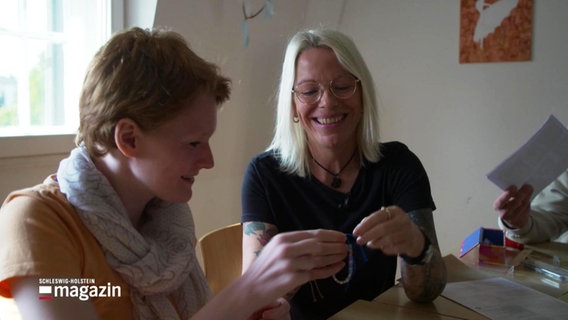 Ergotherapeutin Sonja Rohwer (rechts) sitzt mit einer weiteren Frau am Tisch. Sie lachen und machen Handarbeit. © Screenshot 