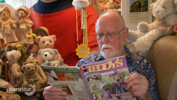 Ein Mann sitzt auf einem Sofa. Er hält eine Zeitschrift ("Teddys") in den Händen. Um ihn herum sind Teddys drappiert. © Screenshot 