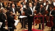 Ein Orchester mit Dirigent steht in einem Konzertsaal. © Screenshot 