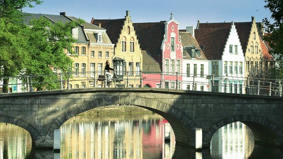 Blick über einen Kanal auf belgische Giebelhäuser in unterschiedlichen Farben. Davor fährt eine Radfahrerin über eine steinerne Brücke. © Screenshot 