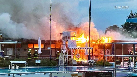 Feuer in einem Freibad. Flammen schlagen aus Gebäuden hinter dem Schwimmbecken. © Screenshot 