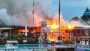 Feuer in einem Freibad. Flammen schlagen aus Gebäuden hinter dem Schwimmbecken. © Screenshot 
