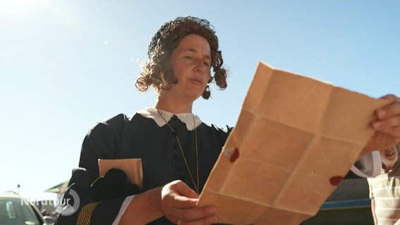 Eine historisch verkleidete Frau liest aus einer Schriftrolle vor. © Screenshot 