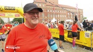 Ein lächelnder Mann mit rotem Shirt und Baseball-Kappe wird interviewt © Screenshot 