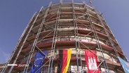 Eine Baustelle eines runden Gebäudes, an dem die europäische, deutsche und Hamburger Flagge hängen. © Screenshot 