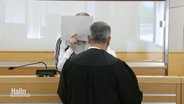 Person mit Robe und Rücken zur Kamera und eine Person dahinter mit Mappe vor dem Gesicht in einem Gerichtssaal. © Screenshot 