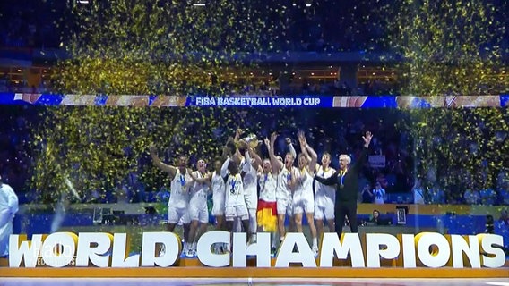 Die Deutsche Basketball-Nationalmannschaft auf einem Podest mit Konfetti und Trophäe. Vor ihr ein Schriftzug: World Champions. © Screenshot 