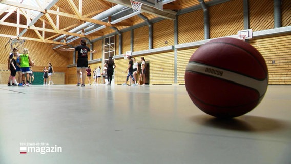 Ein Basketball liegt in einer Turnhalle, im Hintergrund trainieren junge Frauen. © Screenshot 