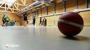Ein Basketball liegt in einer Turnhalle, im Hintergrund trainieren junge Frauen. © Screenshot 