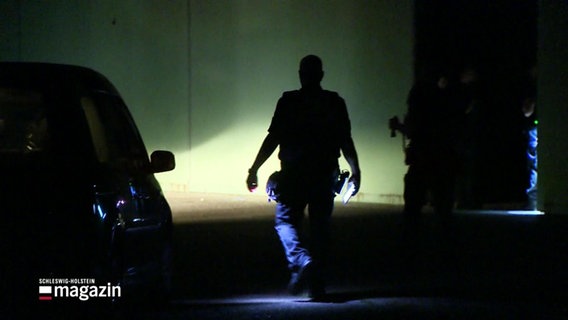 Die Silouette einer Person in Uniform neben einem Polizeiauto vor einer Lagerhalle. © Screenshot 