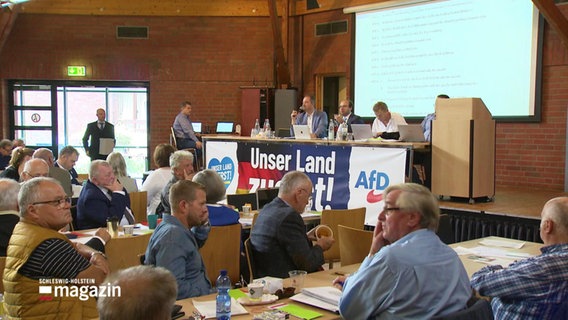 Personen sitzen beim  AfD-Landesparteitag in einem Saal an Tischen. © Screenshot 