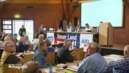 Personen sitzen beim  AfD-Landesparteitag in einem Saal an Tischen. © Screenshot 