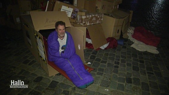 Michael Bahn sitzt, eingehüllt in seinem Schlafsack, in einem offenen Pappkarton. © Screenshot 