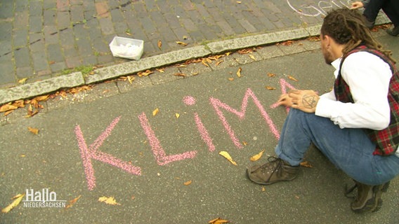 Ein Mann schreibt das Wort "Klima" mit Straßenmalkreide auf eine Straße. © Screenshot 