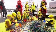 Kita-Kinder in gelben Westen und Bauhelmen stürzen sich mit Schaufeln auf einen großen Berg Lego. © Screenshot 