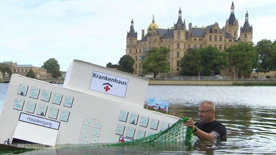Ein Mann und ein halb versunkenes Modell eines Krankenhauses im See vor dem Schweriner Schloss. © Screenshot 